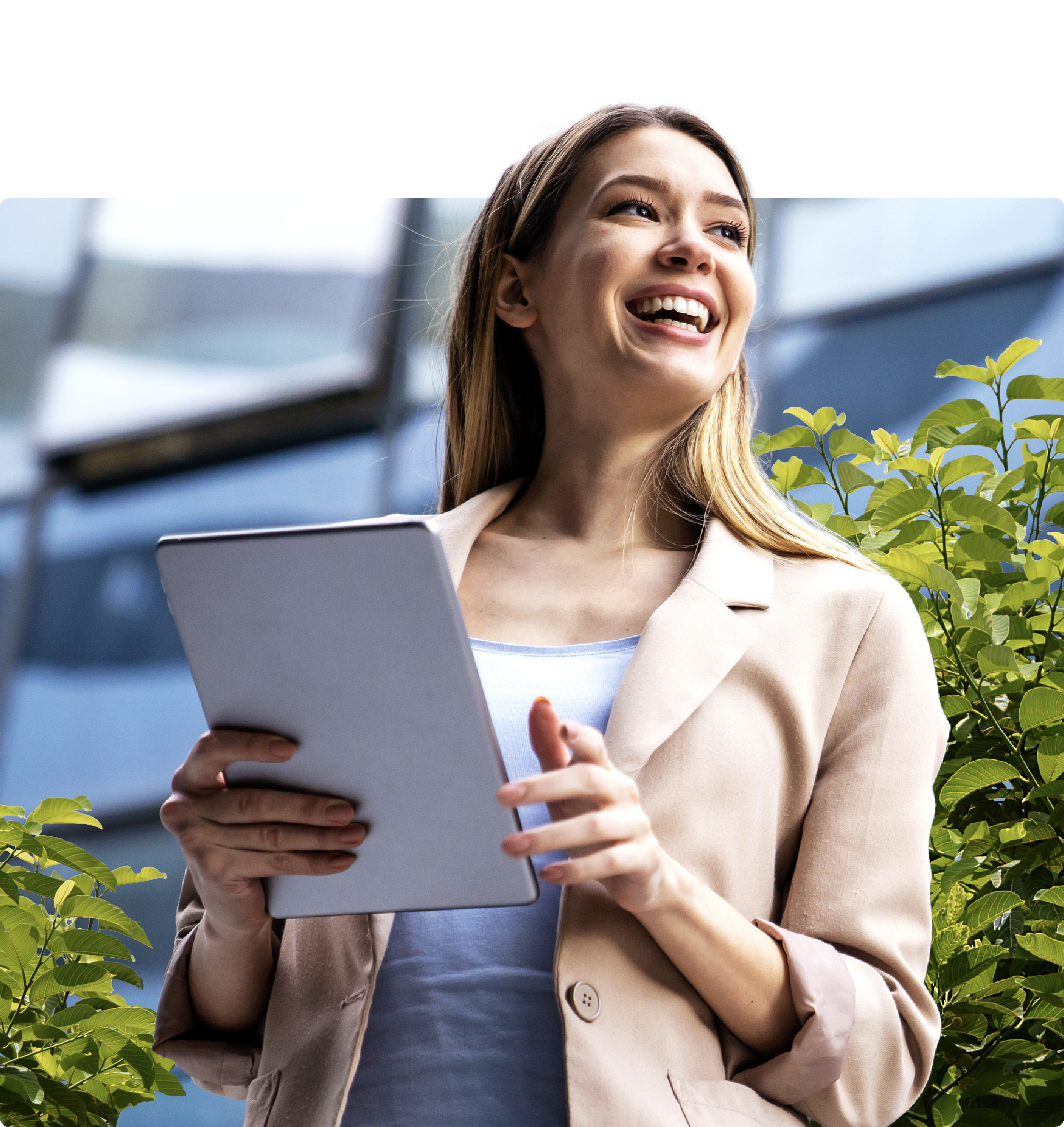 A young businesswoman in a light blazer smiling and holding a tablet while standing outdoors.